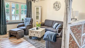 Un salon comprenant un canapé, un fauteuil et un pouf, une petite table avec un vase de fleurs et une horloge au mur.