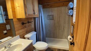 Baño compacto con inodoro, lavamanos y zona de ducha, con paredes de madera y un pequeño detalle de azulejos decorativos.