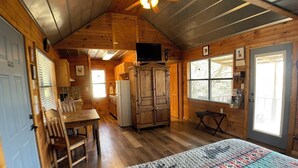 Una habitación acogedora estilo cabaña con interior de madera, una cama con colcha colorida, ventilador de techo, zona de cocina y un ventanal grande.
