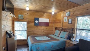 A wooden cabin bedroom with a bed, a chair, a nightstand, and a Texas flag on the wall. - Relaxing Getaway at The Siesta Cabin, Pet Friendly (Fredericksburg)