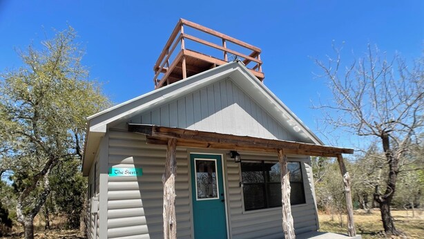 A small cabin with a wooden deck, a green door, and a sign reading "The Siesta". - Relaxing Getaway at The Siesta Cabin, Pet Friendly (Fredericksburg)
