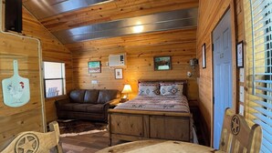 A cozy cabin room with a wooden interior, a bed with patterned bedding, a leather couch, a dining table, and a wooden chair.