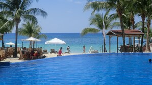 Un área de piscina con sillones de playa y sombrillas, una playa con palmeras y vista al océano.