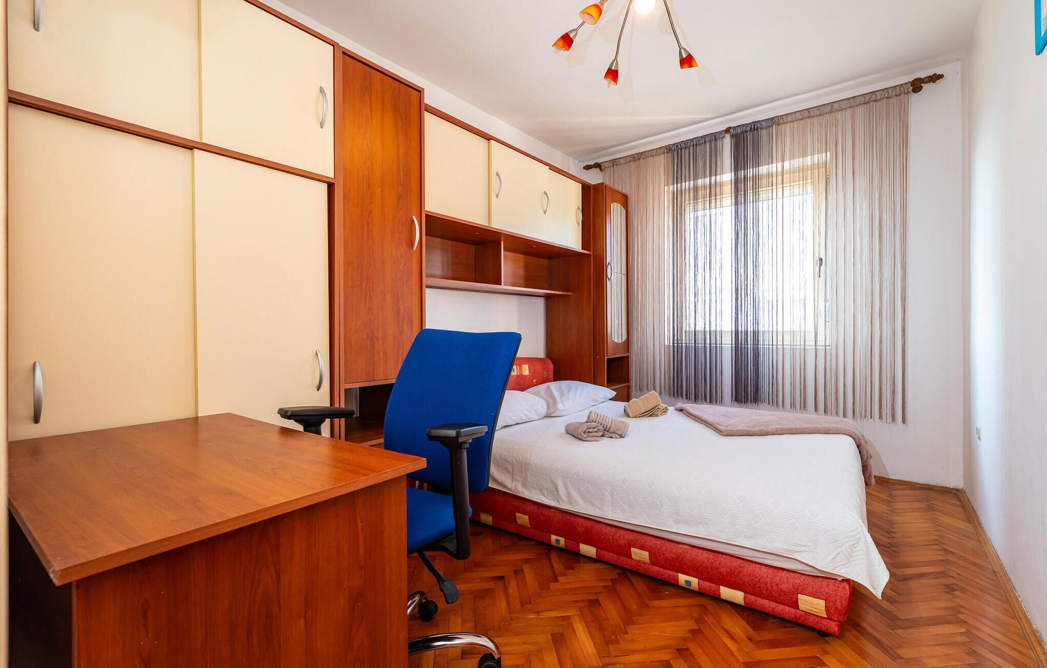 A hotel room with a wooden desk, a blue chair, a bed with a red mattress, and a large window with curtains.