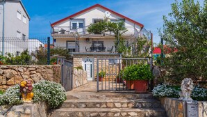 Une maison à deux étages, avec un toit rouge, un balcon et un escalier en pierre menant à l’entrée.