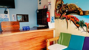 Una zona de recepción con un mostrador de madera, un mostrador de registro, una silla con cojines de colores, un mural en la pared con un jaguar y un extintor.