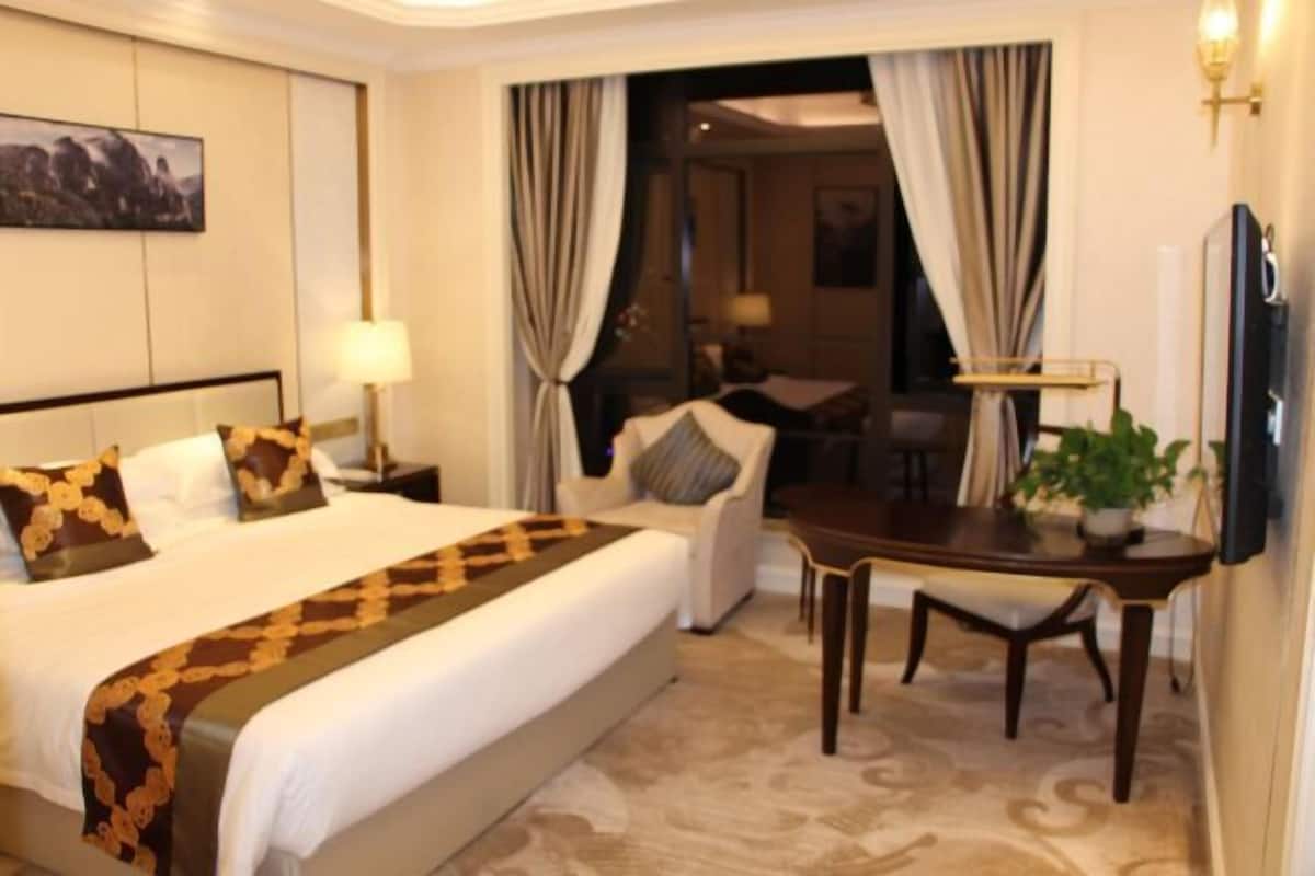 A hotel room with a large bed, a desk, and a chair. There is a lamp on the nightstand and a TV mounted on the wall.