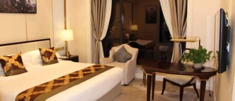 A hotel room with a large bed, a desk, and a chair. There is a lamp on the nightstand and a TV mounted on the wall.