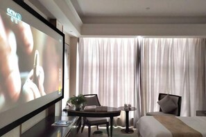 Habitación de hotel con una televisión de pantalla plana grande, una cama, una mesa de comedor con sillas, una mesita con una planta y un ventanal con cortinas.