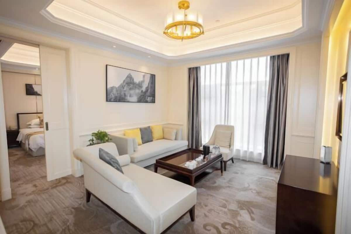 A spacious living room with a white sofa, a coffee table, and a chair. There is a painting on the wall and a large window with curtains.