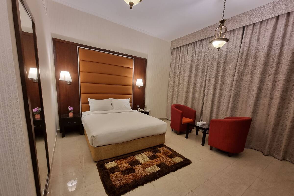 Una habitación de hotel con una cama grande, una mesita de noche con lámpara, un sillón rojo, una pequeña mesa y un ventanal con cortinas.
