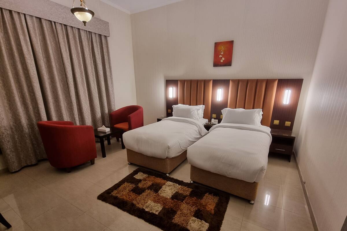 Habitación de hotel con dos camas, un sillón rojo, una mesita, una mesita de noche y una alfombra decorativa.