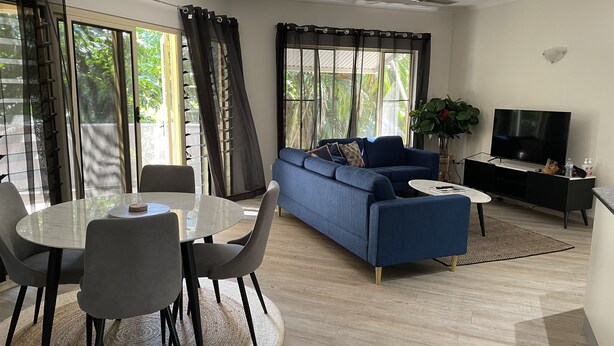 A modern living room with a blue sofa, a dining table with chairs, a TV, and large windows with curtains. - Perfect little home away from home in the tropics. (Parap)