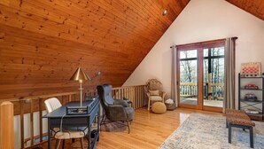 Een gezellig zolderkamer met een schuin houten plafond, een eettafel met stoelen, een bank, een houten bankje en een groot raam met uitzicht op bomen.