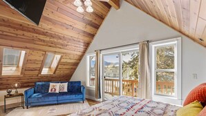 A bedroom with a sloped ceiling, a large bed, a blue sofa, and a view of the outdoors. - Modern Red Lodge Cabin - Walk to Broadway Ave (Red Lodge)