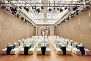 Konferenssisali, jossa on riveissä tuoleja, valaistu lava ja korkea katto.