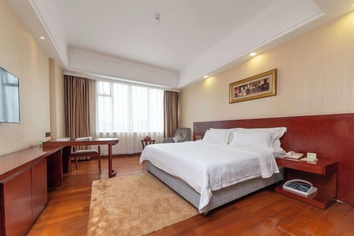 Habitación de hotel con cama, mesita de noche, escritorio, silla y un gran ventanal con cortinas.