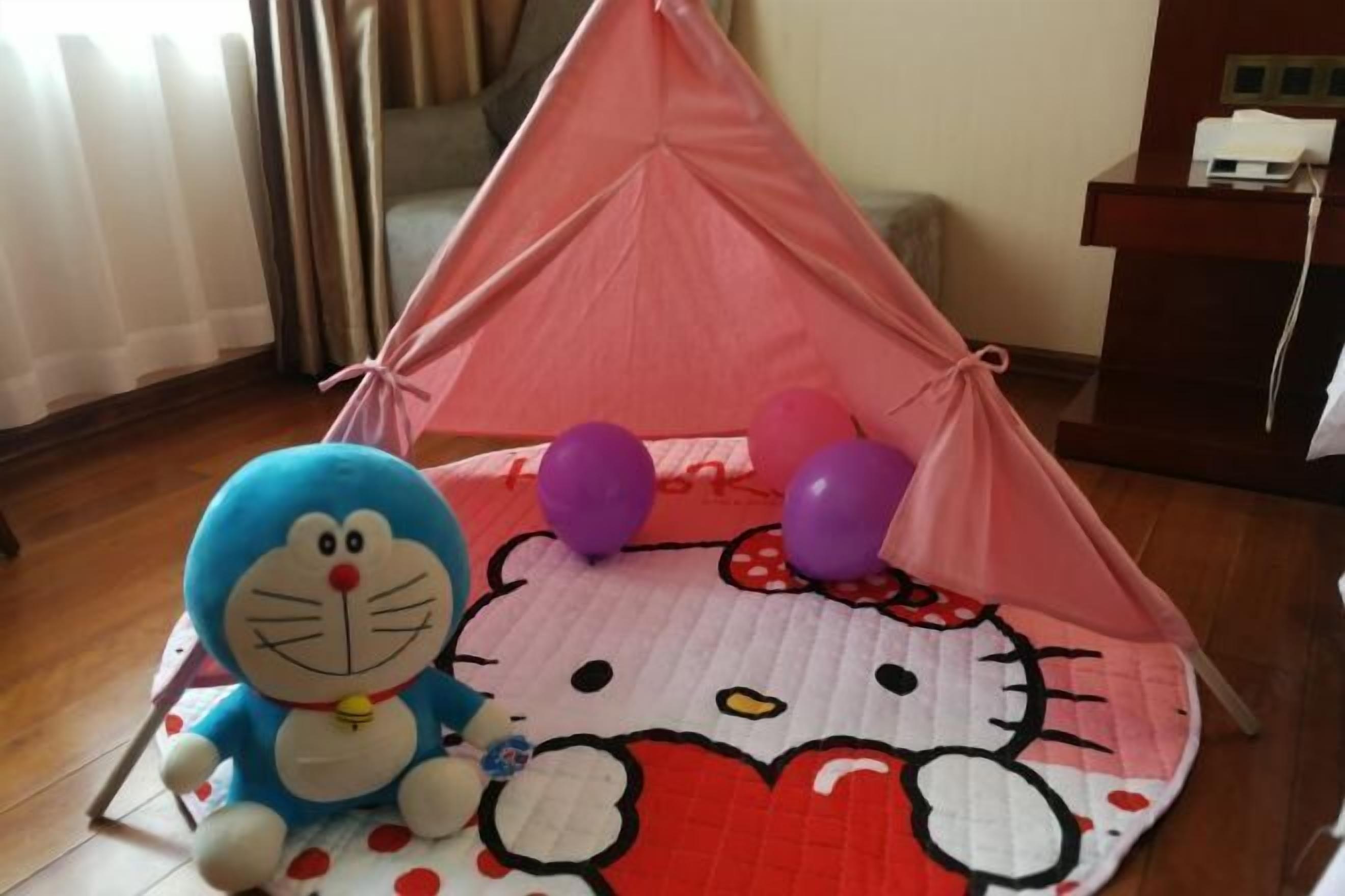 Uma estrutura tipo tenda com design de Hello Kitty, um brinquedo de pelúcia azul e balões roxos no chão.