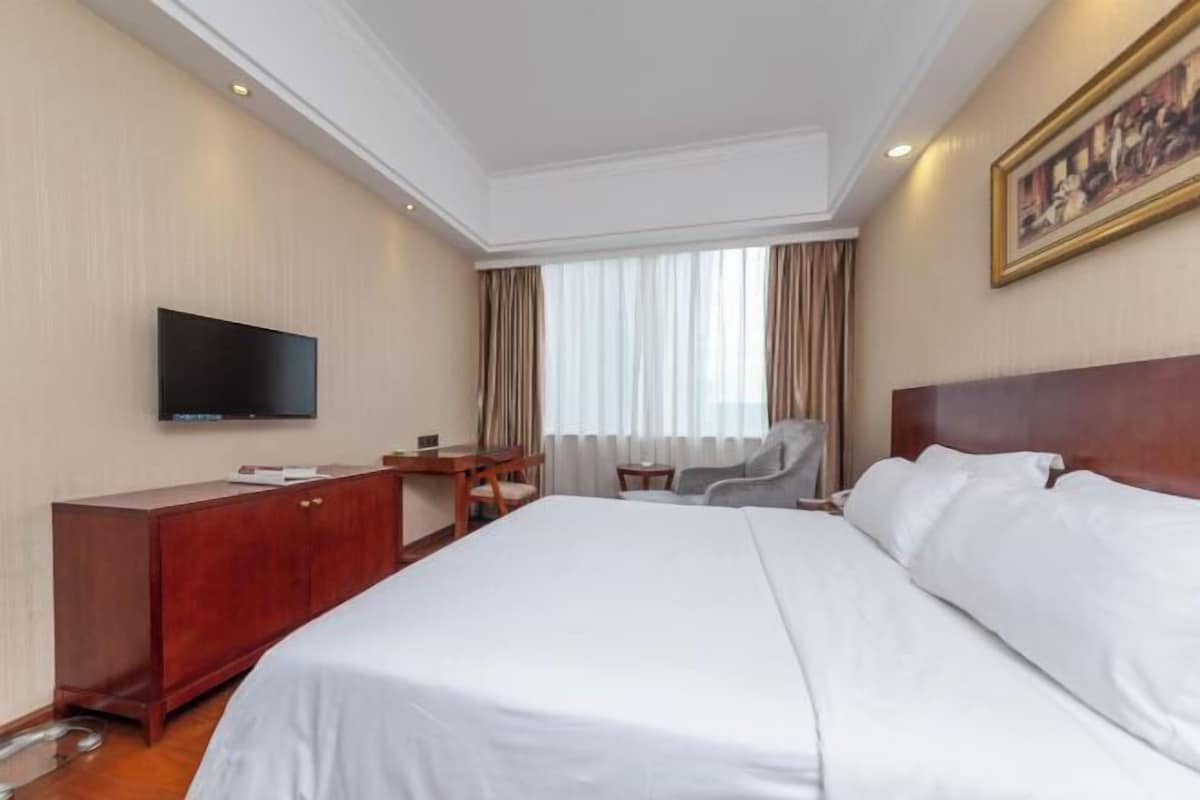 Habitación de hotel con una cama grande, un escritorio, una silla, un televisor y un ventanal con cortinas.