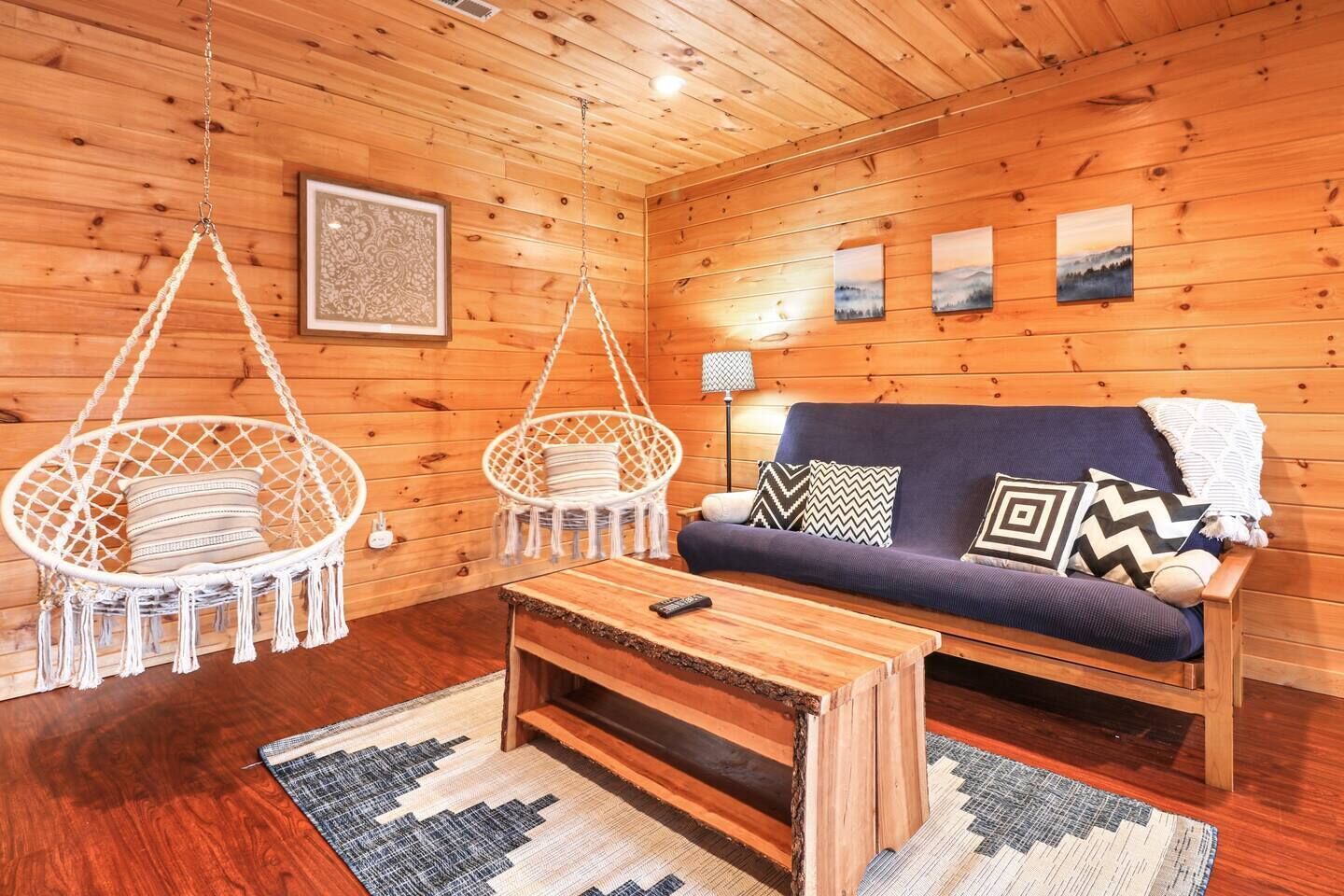A cozy cabin interior with a wooden floor, a sofa with patterned cushions, two hanging chairs, a small table, and a framed picture on the wall.