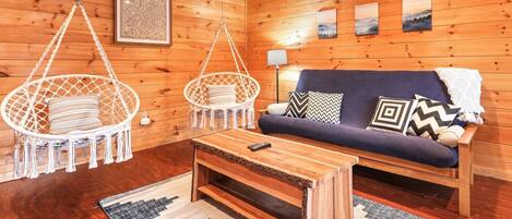 A cozy cabin interior with a wooden floor, a sofa with patterned cushions, two hanging chairs, a small table, and a framed picture on the wall.