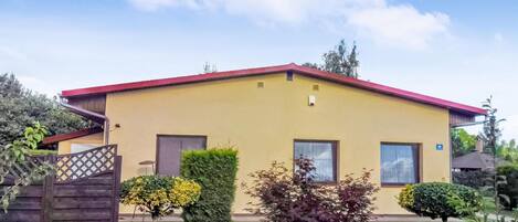 Un edificio amarillo de dos pisos con techo rojo, un pequeño porche y un patio trasero cercado con arbustos variados y un árbol.
