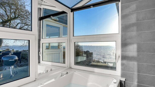 A modern bathroom with a large bathtub, multiple windows offering views of the sea, and a glass-enclosed outdoor area with a grill. - 2 bedroom amazing home in Otterup (Otterup)