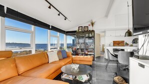 A modern living room with a leather sofa, a coffee table, and a dining area with a white table and chairs. There is a bookshelf filled with books and decorative items, and a large window offering a view of the sea. - 2 bedroom amazing home in Otterup (Otterup)
