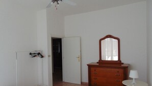 Un dormitorio con una cama, un tocador, un espejo, una mesita de noche con una lámpara y un armario con ropa colgada.