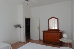 Een slaapkamer met een bed, een dressoir, een spiegel, een klein tafeltje met een lamp en een kast met daarin hangende kleding.