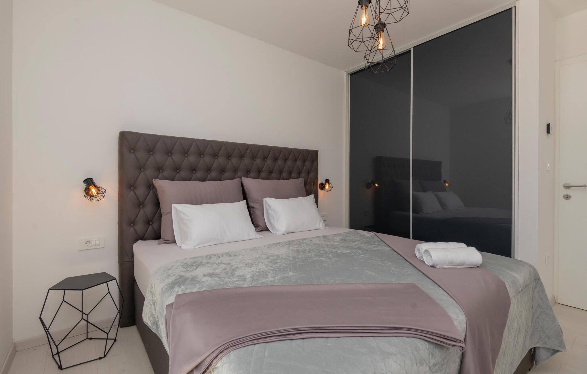 Quarto moderno com cabeceira estofada, cama grande com colcha cinza e rosa, e mesa de cabeceira geométrica.