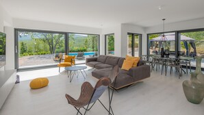 Ett rymligt vardagsrum med en stor grå soffa, en gul sittpuff och en unik fåtölj. Ett matbord med stolar är placerat nära stora fönster som erbjuder utsikt över en pool och grönområden.