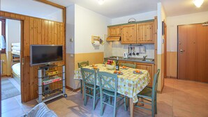 Una cocina compacta con armarios de madera, una mesa de comedor con sillas, un televisor sobre un mueble bajo y un refrigerador pequeño.