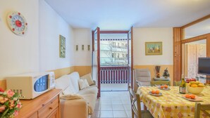 Una habitación acogedora con sofá, una mesa de comedor con fruta, televisor y vistas a edificios a través de una ventana.