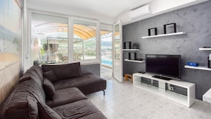 A modern living room with a large sofa, a flat-screen TV, and a balcony with a pool view.
