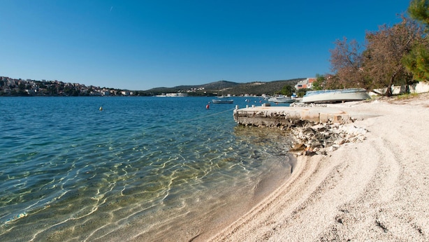 A clear beach with boats docked, a rocky shoreline, and a distant town. - Cozy apartment in Rogoznica with WiFi (Rogoznica)