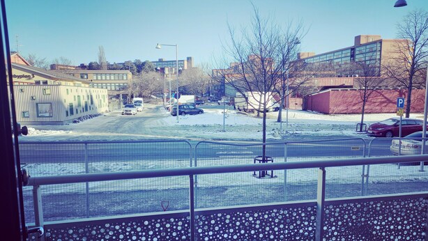 Vista de una calle nevada en la ciudad con edificios, coches y una zona delimitada por una valla.