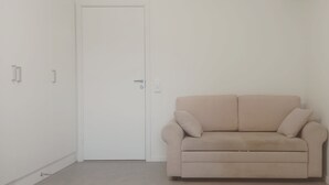 Una habitación minimalista con un sofá color beige, una puerta cerrada y una pared blanca.