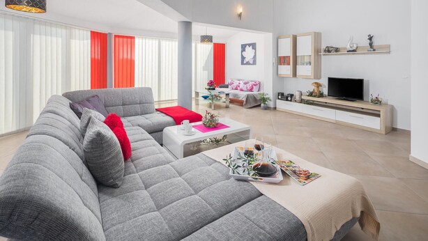 A modern living room with a grey sectional sofa, a coffee table with magazines, a TV on a wooden stand, and a red accent wall. - Lovely home in Valtura (Valtura)