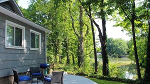 A covered patio with outdoor furniture, a grill, and a view of a lake surrounded by trees. - The Lakeside Cottage in Stow! (Stow)