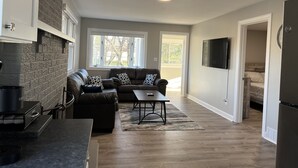 A modern living room with a sectional sofa, a coffee table, and a fireplace. There is a kitchen area with a stove and a refrigerator. A flat-screen TV is mounted on the wall, and there are large windows providing natural light. - The Lakeside Cottage in Stow! (Stow)