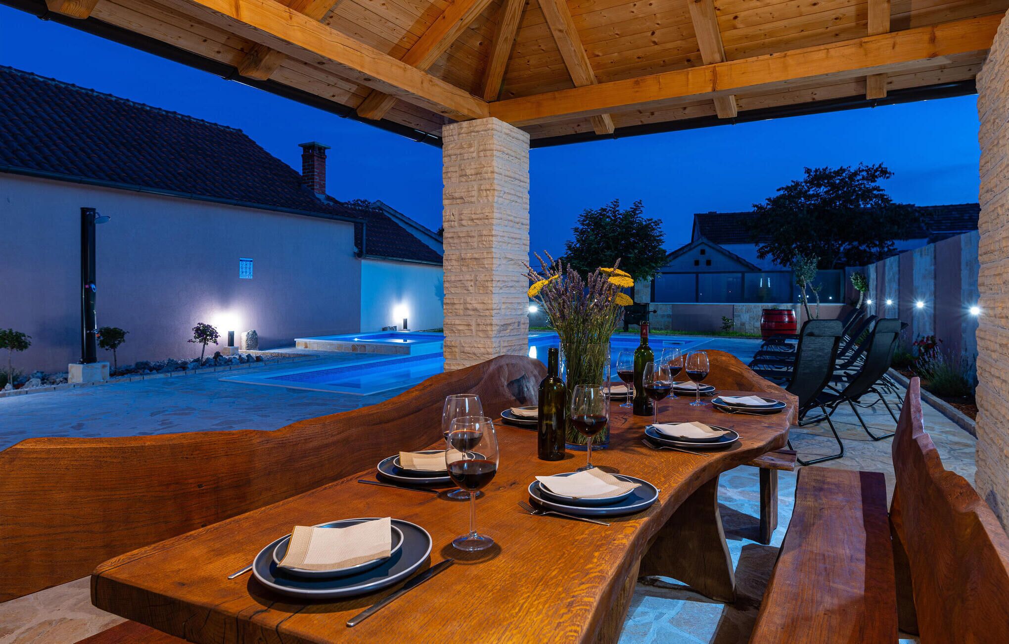 An outdoor dining area with a wooden table set for two, a stone pillar, and a view of a swimming pool and buildings in the background.