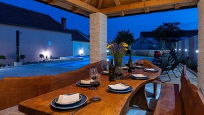 An outdoor dining area with a wooden table set for two, a stone pillar, and a view of a swimming pool and buildings in the background.