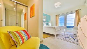 Un sillón amarillo brillante con un cojín colorido, una cama con ropa blanca y almohadas estampadas, un ventanal con cortinas y una vista del cielo.