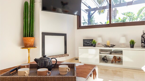 Een gezellige woonkamer met een groot raam, een cactus, een ingelijste foto, een theeservies en een wit kastje met decoratieve spullen.