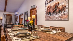 Un comedor con una mesa de madera dispuesta para cuatro personas, rodeada de sillas. Un cuadro grande de un oso adorna la pared, y hay un espejo sobre la puerta.
