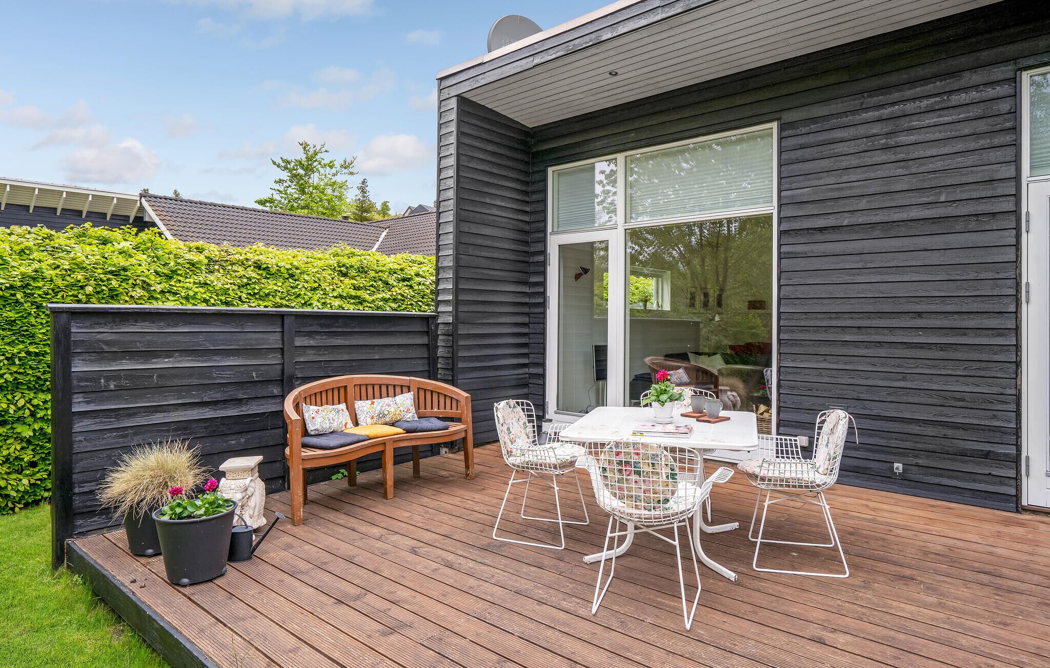 Une terrasse avec un banc en bois et des chaises en fil de fer blanc, une table ornée d’un vase de fleurs et une grande porte vitrée.