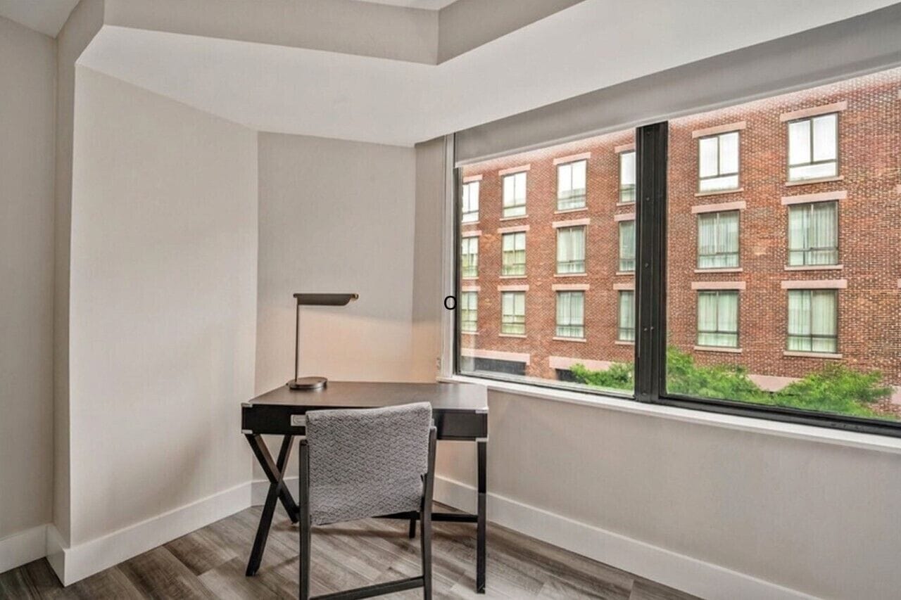A corner room with a desk, chair, and a large window overlooking a brick building.