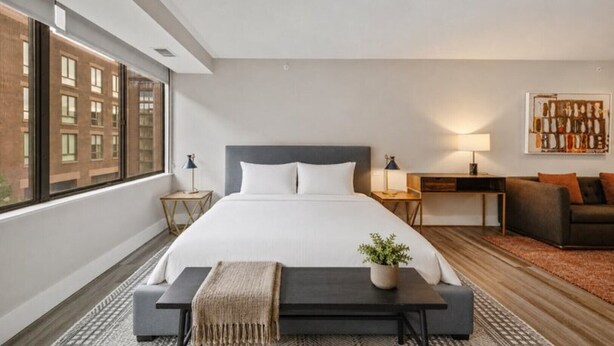 A modern hotel room with a large bed, a sofa, a small table, and a window with a view of buildings. - Global Luxury Suites at Georgetown (Washington)