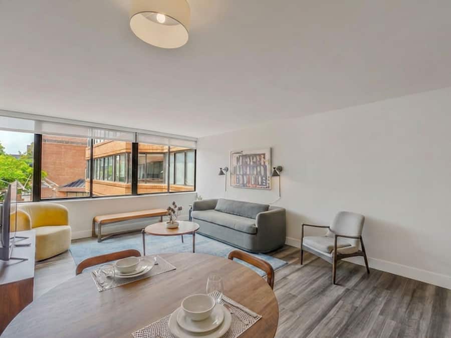 Una sala moderna con comedor, un sofá gris, una mesita de centro y un amplio ventanal con vistas a un edificio de ladrillo.